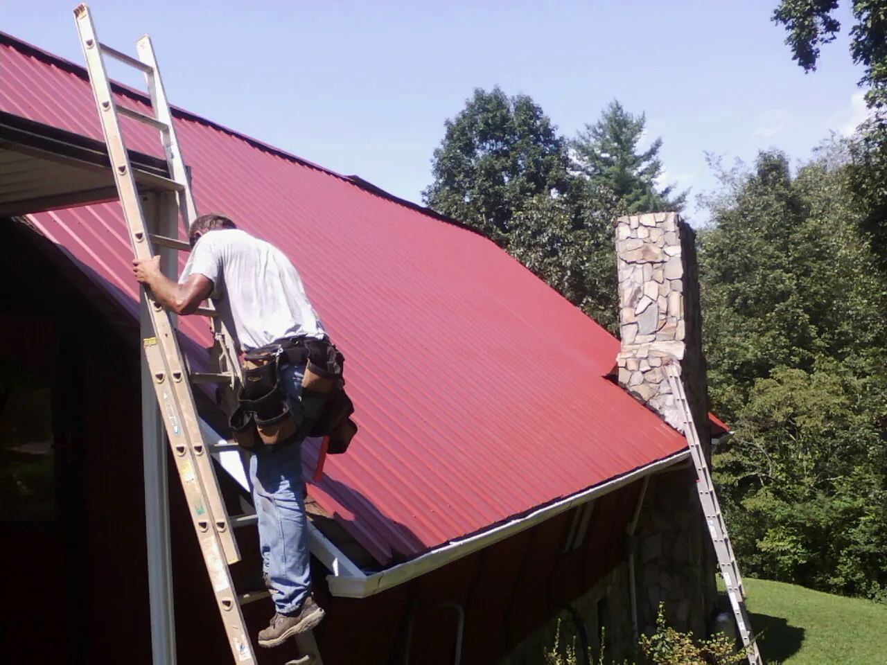 Roofer on a ladder beside a red metal roof and stone chimney — Soffit & Fascia Repair by Roofing Brothers in Robards